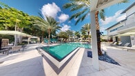 Located at 1640 East Sunrise Boulevard in Fort Lauderdale, Florida, this outdoor amenity space showcases a beautifully designed resort-style pool area that embodies South Florida living at its finest. The centerpiece is a generously sized swimming pool with crystal-clear turquoise water, framed by elegant dark tile borders and surrounded by smooth, light-toned paver decking. Tall, swaying palm trees are planted throughout the area, creating a lush tropical canopy that provides both shade and visual character. A covered pergola structure with a slatted white roof anchors one side of the deck, offering a shaded retreat with outdoor seating and dining arrangements. Rows of contemporary lounge chairs and umbrellas line the pool perimeter, providing ample space for relaxation. Sculptural accents and ornamental plantings add artistic touches to the landscape. The adjacent multi-story building, finished in a clean white modern exterior with green-accented balconies, overlooks the pool deck and reinforces the contemporary architectural aesthetic of the property. The space is bathed in abundant natural Florida sunlight, with a bright blue sky and scattered clouds completing the inviting, resort-like ambiance. This outdoor space serves as a premier leisure destination within the property.