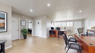 Located at 8150 Cottonwood Ave in Fontana, CA, this single-story office space offers a functional and professional environment designed to accommodate a variety of business needs. The layout is thoughtfully arranged across two distinct areas, beginning with a bright, open reception and main work area. This central space features light wood-style flooring, a textured ceiling with recessed lighting, and multiple warm-toned wooden workstations equipped with computers and guest seating. A wall-mounted HVAC unit and horizontal windows along one wall allow natural light to filter in, creating a welcoming atmosphere for both staff and visitors. A dedicated entryway with a glass-paneled front door and a leasing center welcome mat provides a professional first impression, complemented by potted greenery and a small console table. A private inner office, accessible through a connecting door, features a ceiling fan with light fixture, plantation-shutter windows, built-in closet storage, and a dark wood executive desk with ample workspace. The neutral gray wall tones throughout contribute to a clean, cohesive aesthetic. This office is well-suited for leasing, administrative, or professional service operations, offering a balance of open collaborative space and private work areas.