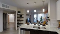 This single-floor space is designed with an open-concept layout that seamlessly connects the kitchen and living areas. The kitchen serves as a central hub, anchored by a large island topped with light granite countertops and a stainless steel double sink. Three sleek pendant lights with frosted glass shades hang above the island, providing focused task lighting while contributing to the modern aesthetic. A stainless steel dishwasher is integrated beneath the counter, adding to the functional design.

Floating dark wood shelves on the adjacent wall offer display space, adding depth and a decorative element to the kitchen zone. Light hardwood-style flooring runs throughout, enhancing the sense of continuity between spaces.

The living area beyond the kitchen is bright and welcoming, benefiting from natural light that streams in through a set of glass doors fitted with horizontal blinds. These doors appear to lead to an outdoor area, extending the sense of space. A television and comfortable seating, including a sofa with accent cushions, furnish the living space. An accent wall in warm terracotta adds a bold contrast to the otherwise neutral color palette, contributing to the overall inviting ambiance.