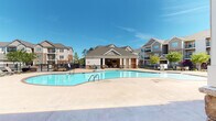 Located at 1300 South Park Way in Sanford, North Carolina, this outdoor amenity space is designed to deliver a resort-style experience for its residents. At the heart of the area is a generously sized, freeform swimming pool with crystal-clear blue water, surrounded by an expansive concrete pool deck that provides ample space for sunbathing and relaxation. A dedicated pergola area with lounge chairs offers additional spots to unwind under the open sky.

The centerpiece of the amenity space is a well-appointed covered pavilion that showcases standout architectural detailing. Substantial stone-clad columns anchor the structure, complemented by a striking full-height natural stone accent wall that serves as a dramatic focal point. The pavilion features a coffered ceiling with ceiling fans to ensure comfortable airflow, and French doors lead into an interior gathering space. Wicker lounge furniture with vibrant yellow cushions provides comfortable outdoor seating arranged around a slatted wood coffee table.

The pavilion opens directly to the pool deck, creating a seamless indoor-outdoor flow. Black wrought-iron fencing encloses the entire pool area, providing security while maintaining an open, airy feel. The surrounding residential buildings, featuring a mix of brick and siding with private balconies, frame the communal space, fostering a strong sense of community in this thoughtfully designed outdoor retreat.