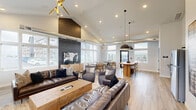 The Union's Community Room in Box Elder, South Dakota, is a thoughtfully designed shared space that seamlessly blends modern aesthetics with warm, inviting functionality. The single-floor layout encompasses a living room, dining area, and kitchen, all unified by light wood-style flooring and a crisp, neutral color palette.

The living area serves as the centerpiece of the room, anchored by a striking floor-to-ceiling tile fireplace with a wood mantel and a wall-mounted flat-screen TV. Rich leather sofas, a chunky wood coffee table, and layered area rugs create a comfortable, lounge-like atmosphere. A dramatic vaulted ceiling with a bold geometric accent wall and a sculptural gold starburst chandelier adds architectural character overhead.

The dining area offers multiple round tables with leather accent chairs, bathed in abundant natural light streaming through large, multi-pane windows. Exposed industrial-style ductwork along the ceiling adds a contemporary edge. A black pendant light and direct outdoor access via a glass-panel door enhance the open, airy feel.

The kitchen is efficiently equipped with stainless steel appliances, dark countertops, gray cabinetry, and floating shelves set against a whitewashed brick backsplash—striking the perfect balance between industrial charm and modern utility. Together, these spaces create a versatile and welcoming community hub.