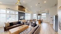 The Union's Community Room in Box Elder, South Dakota, is a thoughtfully designed shared space that seamlessly blends modern aesthetics with warm, inviting functionality. The single-floor layout encompasses a living room, dining area, and kitchen, all unified by light wood-style flooring and a crisp, neutral color palette.

The living area serves as the centerpiece of the room, anchored by a striking floor-to-ceiling tile fireplace with a wood mantel and a wall-mounted flat-screen TV. Rich leather sofas, a chunky wood coffee table, and layered area rugs create a comfortable, lounge-like atmosphere. A dramatic vaulted ceiling with a bold geometric accent wall and a sculptural gold starburst chandelier adds architectural character overhead.

The dining area offers multiple round tables with leather accent chairs, bathed in abundant natural light streaming through large, multi-pane windows. Exposed industrial-style ductwork along the ceiling adds a contemporary edge. A black pendant light and direct outdoor access via a glass-panel door enhance the open, airy feel.

The kitchen is efficiently equipped with stainless steel appliances, dark countertops, gray cabinetry, and floating shelves set against a whitewashed brick backsplash—striking the perfect balance between industrial charm and modern utility. Together, these spaces create a versatile and welcoming community hub.