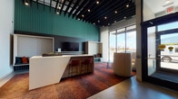 This contemporary residential lobby is thoughtfully designed with a bold, urban aesthetic that makes a strong first impression. The main lounge and reception area features a sleek white quartz counter with rust-colored bar stools, set against a teal accent wall with vertical slat detailing and dark subway tile. A mounted flat-screen display, built-in bench seating with accent pillows, and floor-to-ceiling windows flood the space with natural light while offering open exterior views. Overhead, a dramatic black slatted ceiling with recessed track lighting adds architectural depth and visual interest. The vibrant geometric-patterned carpet anchors the lounge area, contrasting with polished concrete floors throughout the remainder of the lobby. The mailroom area is expansive and open, featuring exposed concrete columns and ductwork, built-in mailboxes, a parcel counter with industrial-style stools, and curated framed artwork. The elevator bank is distinguished by a bold, multicolored triangular-patterned accent wall alongside dark metal wall panels housing multiple stainless steel elevator doors. A fully accessible restroom completes the space, finished with dark ribbed tile wainscoting and an artistic mosaic tile pattern above.