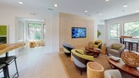 This thoughtfully designed single-floor communal space blends contemporary aesthetics with natural materials to create an inviting and functional environment. The open-plan layout flows seamlessly between a relaxed lounge area and a communal high-top gathering zone, bathed in natural light from large windows and sliding glass doors that frame views of the lush surrounding greenery.

The lounge area is anchored by a striking textured geometric accent wall in warm neutral tones, serving as a dramatic backdrop for a wall-mounted flat-screen display. Curved, modern armchairs in muted grey and chartreuse yellow are arranged around organic wood side tables and natural tree stump accents, lending an earthy warmth to the contemporary setting. A woven area rug grounds the seating arrangement, while recessed lighting illuminates the space from above.

Adjacent to the lounge, a striking live-edge communal table with rich wood grain and clean black metal legs serves as a central gathering point, topped with cascading pendant lights featuring glass globe shades that add an artistic focal point. A slim wood-top counter runs along the windowed wall, offering additional workspace or seating.

Glossy light-toned tile flooring spans the entire space, while accents of vertical moss wall art and diamond-shaped botanical panels add refreshing pops of greenery throughout. Floor-to-ceiling black-framed glass partitions define transitional zones while maintaining the space's airy, open character.