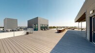 This property features two distinct amenity spaces: a rooftop terrace and a well-appointed fitness center. The rooftop level is defined by a broad, light-toned composite wood deck that extends generously across the top of the building, offering sweeping panoramic views of the surrounding skyline and treeline under open skies. A modern, metal-clad utility structure anchors one end of the deck, featuring large glass doors that connect interior and exterior spaces. The terrace is furnished with a long communal dining table with seating for large groups, as well as lounge chairs positioned for relaxation. Perimeter railings ensure safety while maintaining unobstructed sightlines.

The fitness center is a bright, well-equipped space benefiting from an abundance of natural light through multiple large windows. High ceilings and full-length mirrors enhance the sense of openness. The room is comprehensively outfitted with Precor cardio equipment including treadmills and a rowing machine, a multi-function cable and rack system, a full dumbbell rack, medicine balls, resistance bands, stability balls, and a water rowing machine. Patterned carpet tile flooring adds a contemporary aesthetic, while ceiling fans maintain comfortable airflow throughout the workout area.