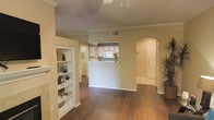 This single-story space showcases a welcoming, open-concept layout that seamlessly connects the main living area to the kitchen. The flooring throughout is a rich, dark hardwood-style material that adds warmth and visual continuity across the space. Crown molding runs along the upper perimeter of the walls, lending an elevated, finished look to the interior.

The living area is highlighted by a built-in shelving unit positioned along one wall, offering both display and storage functionality. A ceiling fan is mounted overhead, contributing to the room's comfort and airflow. Neutral beige wall tones create a clean, versatile backdrop throughout.

The kitchen features white cabinetry and is partially separated from the living area by a half-wall with a pass-through counter, maintaining an open feel while defining the cooking space. Light enters through a window above the kitchen counter, brightening the area. A distinctive arched doorway leads toward what appears to be an entryway, adding an architectural detail that enhances the overall character of the space. The entryway itself is well-lit and features tile flooring, creating a practical transition zone upon entry.