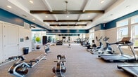 The Aster Fitness Center at 101 McCaw St in Mauldin, SC is a generously sized, single-floor facility designed to support a comprehensive workout experience. The space is defined by its striking architectural ceiling, featuring coffered white panels accented with rich, dark wood beams and recessed lighting, all complemented by ceiling fans that keep the environment comfortable during intense sessions.

The walls are finished in a bold two-tone scheme — a deep teal upper half above crisp white wainscoting — creating a vibrant, energizing atmosphere. Large windows along the perimeter flood the space with abundant natural light while offering views of the surrounding greenery outdoors.

The main workout floor is carpeted and organized into distinct functional zones. A full lineup of cardio equipment, including treadmills, ellipticals, and rowing machines, lines one side of the facility. Opposite, a dedicated strength training area houses a multi-station cable machine, free weights, a dumbbell rack, medicine balls, and adjustable benches. Full-length mirrors along the walls enhance the sense of openness and allow members to monitor their form. A mounted flat-screen TV provides entertainment during workouts. A secondary area near the entrance accommodates additional equipment such as spin bikes and rowing machines, rounding out the facility's versatile offerings.