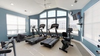 Located at 8101 Fonthill Court in Langhorne, Pennsylvania, this fitness center offers a bright and well-appointed space designed to support an active lifestyle. The room features soaring vaulted ceilings that lend an open, airy feel, complemented by a large arched window arrangement that floods the interior with natural light and provides views of the surrounding neighborhood. The walls are finished in a calming steel-blue tone, creating a focused and energizing atmosphere for workouts. A ceiling fan overhead ensures comfortable air circulation throughout exercise sessions. The fitness center is equipped with a selection of modern cardio machines, including multiple treadmills, an elliptical trainer, and a recumbent stationary bike, all arranged to maximize space and usability. A wall-mounted flat-screen television keeps residents entertained during their workouts, while a wall clock provides convenient time tracking. The flooring is finished with a durable, light-toned surface well-suited for fitness use. Ample natural light from the expansive windows reduces reliance on artificial lighting, enhancing the overall welcoming ambiance of this single-level amenity space.