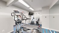 This single-level fitness center is designed to support a well-rounded workout experience, offering a thoughtfully arranged collection of cardio and strength training equipment. The space is divided into two connected zones, each serving a distinct function while maintaining a cohesive aesthetic throughout.

The cardio area features multiple treadmills, elliptical trainers, and a recumbent stationary bike, all positioned on a patterned carpet with bold blue and gray striping. A wall-mounted flat-screen television provides entertainment during workouts, while frosted glass panels and a window allow natural light to filter into the room. Overhead fluorescent and skylight-style fixtures ensure consistent, bright illumination.

The strength training zone is lined with full-wall mirrors that visually expand the space and allow users to monitor their form. A multi-function cable machine and additional resistance equipment are arranged along the mirrored wall, which is further accented with blue diagonal design elements.

A small amenity alcove houses a sanitizing station, waste receptacles, and informational displays. An accessible, ADA-compliant restroom is conveniently located within the facility. The overall design is clean and modern, with white walls, bright lighting, and a sporty color palette creating an energizing atmosphere.