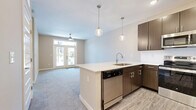 Located in Mason, OH, this one-bedroom, one-bathroom unit offers a thoughtfully designed open-concept layout that maximizes its efficiently used floor space. The combined kitchen and living area serves as the heart of the home, where the kitchen stands out with rich, dark-toned cabinetry, white quartz countertops, and a full suite of stainless steel appliances including a range, microwave, refrigerator, and dishwasher. A peninsula island with an undermount sink provides additional prep space and casual seating potential, while two globe pendant lights add a modern, decorative touch overhead. Light wood-look flooring runs throughout the kitchen, transitioning seamlessly into plush carpeting in the living area. French doors at the far end of the living room flood the space with natural light and provide outdoor access, complemented by a ceiling fan for year-round comfort. The bedroom is a comfortable retreat, featuring soft carpeting, a ceiling fan, and a window with views of the surrounding community. A dedicated walk-in closet adjoins the bedroom, offering generous storage. The unit combines contemporary finishes with practical design, creating a clean, modern living environment well suited for everyday living.