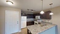 This single-floor residence in Burlington, NC, along the I-40 corridor offers a practical and cohesive layout designed for comfortable everyday living. The open-concept design connects the living area directly to the kitchen, creating a spacious, unified flow throughout the main living space. Neutral-toned carpet covers the living and bedroom areas, complementing the warm beige walls found throughout the unit.

The kitchen is a standout feature, outfitted with rich dark espresso cabinetry that extends to the ceiling, providing generous storage. Granite-look countertops line the workspace, and a peninsula with pendant lighting serves as a natural divider between the kitchen and living area. Wood-look flooring, a white refrigerator, electric range, and built-in microwave complete the functional cooking space. Track lighting overhead ensures the kitchen is well-illuminated.

The bedrooms are generously sized, with one featuring a ceiling fan, a window with exterior views, and access to a closet. Additional bedrooms offer natural light through blinds-equipped windows. The bathroom includes a full tub and shower combination with white tile surround and wood-look flooring, maintaining a clean, uniform aesthetic throughout the home.
