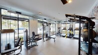 This contemporary fitness center is thoughtfully designed to support a full range of workout routines in a bright, welcoming environment. The space is defined by expansive floor-to-ceiling windows along multiple walls, flooding the interior with natural light and providing open views of the surrounding outdoor greenery. Exposed silver ductwork runs along the upper portion of the walls, lending an industrial character that complements the clean, modern aesthetic.

The strength training area is well-appointed with a multi-station cable machine, a dedicated leg press machine, a flat weight bench, and a full dumbbell rack spanning a wide range of weights. A large wall-mounted mirror in the free weights section enhances the sense of space and allows users to monitor their form during workouts. Durable rubber-composite flooring throughout provides safety and comfort underfoot.

The cardio zone features multiple treadmills, an elliptical trainer, a stationary bike, and TRX suspension training straps, offering diverse options for cardiovascular and functional fitness training. A ceiling fan aids in air circulation during intense sessions. Glass-panel entry doors connect the fitness center to the adjacent facility, ensuring convenient access. Overall, the space delivers a professional gym experience in a clean, modern, and naturally lit setting.