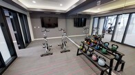 This dedicated fitness studio is thoughtfully designed to support a wide range of workout routines in a sleek, modern environment. The space features a contemporary aesthetic with dark gray accent walls, a tray ceiling with recessed lighting, and a neutral-toned flooring accented with subtle striped detailing that adds visual energy to the room. Multiple wall-mounted flat-screen TVs are positioned for easy viewing during workouts, enhancing the exercise experience. A full floor-to-ceiling mirror runs along one wall, visually expanding the space while allowing users to monitor their form. The studio is equipped with a row of spin bikes arranged for group or individual cycling sessions, alongside a well-stocked tiered kettlebell rack offering a broad range of weights in various colors for strength training and functional fitness. The layout is open and uncluttered, providing ample room to move freely between equipment. The combination of smart lighting, mirrored walls, and purpose-built equipment makes this fitness studio a highly functional and motivating space for residents or guests seeking a convenient, professional-grade workout environment.