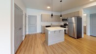 This single-floor residence presents a cohesive, contemporary design throughout. The kitchen serves as a central highlight, outfitted with a full suite of stainless steel appliances, including an electric range with overhead microwave, dishwasher, and refrigerator. Light-toned flat-front cabinetry extends to the upper walls, providing generous storage, while clean white countertops offer ample workspace. A central island with an integrated breakfast bar adds additional prep space and casual seating, illuminated by sleek pendant lighting overhead. Light hardwood-style flooring runs seamlessly throughout the unit, enhancing the bright, airy atmosphere.

The open layout connects the kitchen directly to the living area, which benefits from a large sliding glass door that invites natural light and provides access to an outdoor space. The living area is spacious and versatile, with clean white walls and recessed lighting contributing to a fresh, modern feel.

The bathroom maintains the same contemporary aesthetic, featuring a light wood vanity with a white countertop, an undermount sink, chrome fixtures, a large mirror, and built-in drawer storage. The overall design of the space is clean, functional, and consistent in its modern, neutral palette.