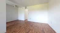 This single-floor unit is laid out to maximize functionality across its key living spaces. The living area is open and airy, featuring warm-toned hardwood-style flooring that flows consistently throughout the unit. A ceiling fan is mounted overhead, and a wall-mounted air conditioning unit provides climate control. Large sliding glass doors in one room open to an outdoor view of lush greenery, allowing natural light to fill the space. A half-wall partition separates the living area from the kitchen, which features white cabinetry, granite-style countertops, a textured tile backsplash, and a freestanding electric range. A security screen door in the kitchen offers additional ventilation and outdoor access. The bedroom is generously sized with clean white walls, hardwood-style flooring, and access to a closet. The bathroom is a standout feature, showcasing bold mint green and white vertically striped ceramic tile work surrounding a full-size soaking tub with an overhead shower. The overall aesthetic of the unit combines retro character with practical, easy-to-maintain finishes throughout.