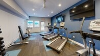 Located at 1712 Pinecroft Dr in Taylors, SC, this fitness center is a thoughtfully designed, single-room facility built to support a well-rounded workout routine. The space features a modern aesthetic with bold blue accent walls, recessed lighting, and a patterned carpet tile floor in alternating shades of teal and brown that adds visual energy to the room.

A striking full-wall photographic mural dominates one end of the space, depicting dynamic athletic imagery — including a runner, cyclist, and yoga practitioner — rendered in black and white for a dramatic, motivational effect. This design detail sets a strong tone for the room's purpose.

The cardio section is anchored by a row of TRUE brand treadmills and a stationary spin bike, all positioned toward the windowed wall to allow natural light and outdoor views of a surrounding patio and wooded landscape. A wall-mounted flat-screen television is positioned above the cardio equipment for entertainment during workouts.

Strength training is supported by a multi-function cable machine and a full dumbbell rack stocked with a wide range of free weights, providing ample options for resistance training. A ceiling fan and recessed lighting ensure the room remains well-ventilated and well-lit throughout. A glass door leads directly to an outdoor patio area, further enhancing the open, accessible feel of this practical and inviting fitness space.