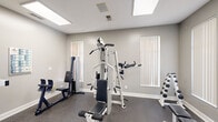 Located at 5701 Ebner Dr in Dublin, Ohio, this fitness center offers a thoughtfully designed workout environment spread across a single floor. The space is divided into two functional zones, each catering to different aspects of physical training.

The cardio area is bright and inviting, featuring multiple treadmills positioned in front of large windows that provide natural light and views of the adjacent pool area. A wall-mounted television and a full-length mirror add practicality and a sense of openness to this zone, while the rubberized flooring ensures comfort and safety during workouts.

The strength training area is equally well-appointed, housing a multi-station cable machine, a lat pulldown unit, a dumbbell rack, and stability balls, making it suitable for a full-body resistance routine. An exercise chart on the wall and ample open floor space support a variety of training styles. Overhead skylights brighten the room naturally throughout the day.

A welcoming entry area with neutral gray walls, polished aggregate flooring, and a decorative console table provides a clean transition into the workout space. A restroom is conveniently accessible from within the facility. The overall aesthetic is clean, modern, and functional, with a neutral color palette and consistent flooring throughout that creates a cohesive, professional atmosphere.