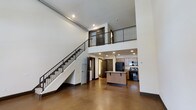 Unit 1303 is a dramatic multi-level loft spanning three floors, designed to impress with its soaring double-height ceilings and open-concept layout. The main living area is anchored by polished concrete floors that lend an industrial yet refined character to the space. A sleek staircase with dark metal railings rises prominently along one wall, connecting the levels while serving as a striking architectural focal point. Recessed lighting throughout ensures the space is well-lit, complementing the airy, voluminous feel created by the expansive vertical height. The kitchen is seamlessly integrated into the open floor plan, featuring warm wood cabinetry, a central island with bar seating, granite countertops, and black appliances, including a gas range and refrigerator. An upper-level mezzanine overlooks the main living area below, framed by matching metal railings that echo the industrial aesthetic seen throughout. The clean white walls contrast beautifully with the dark metal accents and warm wood tones, creating a balanced and cohesive design. With multiple levels, generous vertical space, and thoughtfully curated finishes, this loft offers a distinctive urban living experience that is both functional and architecturally compelling.
