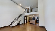 Unit 1303 is a dramatic multi-level loft spanning three floors, designed to impress with its soaring double-height ceilings and open-concept layout. The main living area is anchored by polished concrete floors that lend an industrial yet refined character to the space. A sleek staircase with dark metal railings rises prominently along one wall, connecting the levels while serving as a striking architectural focal point. Recessed lighting throughout ensures the space is well-lit, complementing the airy, voluminous feel created by the expansive vertical height. The kitchen is seamlessly integrated into the open floor plan, featuring warm wood cabinetry, a central island with bar seating, granite countertops, and black appliances, including a gas range and refrigerator. An upper-level mezzanine overlooks the main living area below, framed by matching metal railings that echo the industrial aesthetic seen throughout. The clean white walls contrast beautifully with the dark metal accents and warm wood tones, creating a balanced and cohesive design. With multiple levels, generous vertical space, and thoughtfully curated finishes, this loft offers a distinctive urban living experience that is both functional and architecturally compelling.