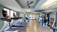 This fitness center offers a clean, well-appointed environment designed to support a full range of workout routines. The space is anchored by warm hardwood flooring that runs throughout, providing a polished and professional atmosphere. A row of treadmills lines one side of the room, complemented by elliptical machines and a variety of strength and resistance training equipment, ensuring comprehensive fitness options for users of all levels.

The architectural design stands out with elegant coffered ceilings finished in a soothing teal-blue accent color, trimmed with crisp white molding. Ceiling fans are mounted within the coffers, providing consistent air circulation throughout the space. Large windows along the perimeter flood the room with natural light, creating an open and energizing ambiance.

A glass-paneled partition wall at the rear of the fitness center adds a sense of depth and visual openness, while also separating distinct workout zones. The overall layout is thoughtfully organized, allowing for comfortable movement between equipment. The combination of stylish design elements and practical amenities makes this fitness center both functional and inviting for daily use.