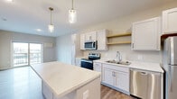 This efficiently designed one-bedroom, one-bathroom unit makes excellent use of its single-floor layout, offering a cohesive and functional living environment. The open-concept kitchen and living area serves as the heart of the unit, featuring white shaker-style cabinetry with matte black hardware, light quartz countertops, a central island ideal for dining or meal prep, and a full suite of stainless steel appliances. Warm hardwood-style flooring runs throughout the main living spaces, while two stylish glass pendant lights with brass accents illuminate the kitchen. A dedicated teal entry door adds a distinctive design touch upon arrival. The living area connects seamlessly to a private balcony through sliding glass doors, offering open outdoor views. The bedroom is carpeted for comfort and features a large window that brings in natural light along with views of the surrounding area. A dedicated in-unit laundry space houses a full-size top-load washer and dryer. The bathroom offers a clean, functional design with a wide vanity, ample storage drawers, wood-look flooring, and a tub-shower combination. Overall, the unit presents a modern, neutral palette with thoughtful finishes suited for comfortable everyday living.