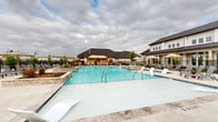 This single-story community amenity facility is designed to offer a well-rounded outdoor living experience. At its centerpiece is a generously sized resort-style swimming pool with a gradually sloping entry, framed by travertine-style pool decking, stone-accented raised planters, and an array of lounge chairs and market umbrellas that create a relaxed, resort-inspired atmosphere.

Adjacent to the pool is a spacious covered pavilion that serves multiple functions. The structure features soaring vaulted ceilings fitted with ceiling fans, ensuring comfortable airflow throughout. Large open-air bays allow natural light to flood the interior while offering unobstructed views of the surrounding community grounds. Within the pavilion, a substantial dining table with lattice-back chairs accommodates large gatherings, while a separate lounge area with upholstered seating and a concrete bowl coffee table provides a quieter retreat.

An extended open-air corridor runs through the facility, supported by clean white columns and covered ceilings with additional ceiling fans overhead. This passageway connects the various zones of the amenity space and opens to the landscaped community grounds on either end. The overall design emphasizes an indoor-outdoor connection, with a neutral, modern aesthetic throughout.