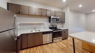 This single-floor unit offers a clean, contemporary design with a cohesive aesthetic carried throughout each room. The kitchen serves as a functional centerpiece, showcasing rich dark-toned cabinetry that extends to the ceiling, stainless steel appliances including a refrigerator, range, microwave, and dishwasher, and light granite-style countertops. A white subway tile backsplash adds a modern touch, while a central island provides additional prep space and casual seating. Warm, light-toned hardwood flooring runs throughout the main living areas, contributing to a bright and welcoming atmosphere.

The bedroom is a calm, neutral space featuring plush wall-to-wall carpeting for added comfort. A large window floods the room with natural light and offers views of the surrounding area, making the space feel open and airy.

The bathroom is well-equipped with a soaking tub and shower combination, a wide vanity with dark wood cabinetry and ample drawer storage, a large mirror with overhead lighting, and a matching tall storage cabinet. The continuation of hardwood flooring into the bathroom ties the space together seamlessly.

Overall, this unit presents a thoughtfully designed interior that balances modern style with practical functionality, making it a comfortable and move-in-ready space.