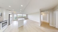 This single-floor unit offers a clean, contemporary design throughout, finished with light hardwood-style flooring and neutral white walls that create a bright, cohesive aesthetic. The open-concept kitchen and living area forms the heart of the unit, where a sleek kitchen island with a white quartz countertop serves as a central feature. The kitchen is equipped with high-gloss white cabinetry extending to the ceiling, stainless steel appliances including a refrigerator, range, microwave, and dishwasher, and recessed lighting overhead. The adjoining living area is generously proportioned, bathed in natural light from a wide bank of windows that frame outdoor views of the surrounding landscape and open sky.

Four bedrooms are accessible via a connecting hallway, each featuring the same light flooring, neutral walls, and windows with baseboard heating beneath. The primary bedroom offers a comfortable footprint suitable for a full bedroom suite. The single full bathroom is finished in white tile, featuring a vanity with an integrated sink, mirrored medicine cabinet, brushed nickel fixtures, and a tub-shower combination. The overall ambiance is clean, minimalist, and light-filled throughout.