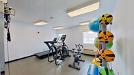 This single-floor fitness room is designed to support a comprehensive workout experience, offering a diverse selection of equipment within a clean, functional layout. The space is finished with neutral white walls, dark base trim, and durable rubber-floored exercise zones, creating a practical and focused environment for physical training.

Cardio equipment includes multiple treadmills and an elliptical trainer, positioned near a window that allows natural light to enter the room. A stationary spin bike provides an additional lower-impact cardiovascular option. The room also features a Matrix dual-cable functional trainer, enabling a wide range of strength and resistance exercises.

Accessories are well-organized throughout the space. A dedicated rack holds an assortment of colorful medicine balls and resistance bands, while stability balls in various sizes are mounted on a wall-mounted storage rack. Exercise mats are also available for floor-based workouts.

Overhead, flush-mounted fluorescent panel lights ensure consistent, even illumination across the entire room. A small utility area with cabinetry and a sink is visible in one corner, adding a convenient practical element. The overall ambiance is clean, organized, and purpose-driven, suited for both light and moderate fitness routines.