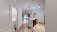 Located at 1100 South Blvd in Charlotte's vibrant South End neighborhood, this one-bedroom, one-bathroom unit offers a well-designed open-concept layout across a single floor. The combined living, dining, and kitchen area serves as the heart of the home, anchored by a curved granite-topped island with an undermount sink and elegant pendant lighting overhead. The kitchen is outfitted with rich dark cabinetry, granite countertops, a subway tile backsplash, and a full suite of stainless steel appliances including a refrigerator, range, and microwave. Light wood-tone flooring runs throughout the main living areas, complemented by ceiling fans and a wall of French doors that invite natural light into the living room. The bedroom features plush carpet, a ceiling fan, and a window with outdoor views, along with access to a spacious walk-in closet equipped with wire shelving. The bathroom includes a tiled tub-shower combination, a dark-cabinet vanity with a granite countertop, and matching wood-tone flooring. A dedicated laundry area accommodates a full-sized washer. Additional closet and utility spaces round out this efficiently planned and thoughtfully finished home.