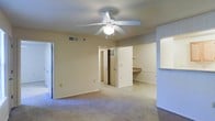 This single-floor space presents an open and functional layout highlighted by neutral cream-toned walls and light carpeting that contribute to a clean, understated aesthetic. The main living area is centrally anchored by a white ceiling fan with an integrated light fixture, providing both air circulation and ambient lighting. HVAC vents are visibly integrated into the ceiling, ensuring year-round climate control throughout the space.

Toward the rear of the main area, a built-in shelving unit with a fixed countertop surface offers practical storage and display options. Adjacent to this, a pass-through window or opening connects to a neighboring room, enhancing the sense of openness and allowing for easy communication between spaces. The open doorways visible in the background suggest additional rooms, including what appears to be a closet with louvered doors, providing ample storage.

The transition between carpet and hard flooring near the rear of the space indicates distinct functional zones within the layout. The overall design is straightforward and versatile, offering a blank canvas well-suited for a variety of uses or furnishing arrangements.