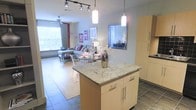 This single-floor space showcases an open-concept layout that integrates the kitchen, dining, and living areas into one cohesive environment. The kitchen is anchored by a central island topped with light granite countertops, complemented by light wood cabinetry with sleek chrome hardware. Pendant lights hang above the island, while a track lighting system overhead provides additional illumination throughout the space.

The flooring transitions from dark tile in the kitchen zone to warm hardwood-style flooring in the dining and living areas, visually defining each functional zone while maintaining an open flow. A small dining area with transparent acrylic chairs sits adjacent to the kitchen, adding a modern, airy aesthetic.

The living area extends toward a large window that invites abundant natural light, offering views of the surrounding greenery. A ceiling fan is positioned above the seating area, contributing to comfort. A wall-mounted mirror in the transition zone adds depth and reflects light throughout. Framed wall art and neutral gray walls complete the contemporary, well-lit ambiance of the space.