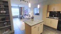 This single-floor space showcases an open-concept layout that integrates the kitchen, dining, and living areas into one cohesive environment. The kitchen is anchored by a central island topped with light granite countertops, complemented by light wood cabinetry with sleek chrome hardware. Pendant lights hang above the island, while a track lighting system overhead provides additional illumination throughout the space.

The flooring transitions from dark tile in the kitchen zone to warm hardwood-style flooring in the dining and living areas, visually defining each functional zone while maintaining an open flow. A small dining area with transparent acrylic chairs sits adjacent to the kitchen, adding a modern, airy aesthetic.

The living area extends toward a large window that invites abundant natural light, offering views of the surrounding greenery. A ceiling fan is positioned above the seating area, contributing to comfort. A wall-mounted mirror in the transition zone adds depth and reflects light throughout. Framed wall art and neutral gray walls complete the contemporary, well-lit ambiance of the space.