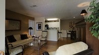 This single-floor space is thoughtfully designed with an open-concept layout that seamlessly connects the living, kitchen, and dining areas. The living area anchors the space with a cream-toned sofa, a zebra-print accent chair, and a glass-top coffee table, creating a stylish yet functional seating arrangement. Warm hardwood flooring extends throughout, tying each zone together cohesively. The kitchen features white upper cabinetry, a peninsula with bar-height stools, and dark appliances, including a refrigerator. The peninsula serves as both a practical workspace and a casual dining counter. Adjacent to the kitchen, a dedicated dining area accommodates a dark-toned table and chairs, suitable for more formal meals. The walls are finished in warm taupe tones, complemented by white trim and wainscoting details near the dining zone. A textured ceiling with recessed and flush-mounted lighting provides even illumination throughout. Framed wall art and a tall table lamp with a black shade add decorative character to the living area. A doorway visible near the living space suggests additional rooms beyond the main open-plan zone.