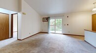 Located on Green Road in Pewaukee, Wisconsin, this two-bedroom, two-bathroom two-story residence offers a practical and comfortable layout across two levels. The main living area on the upper floor features soft carpeting and vaulted ceilings that lend an open, airy feel. A sliding glass door opens to a private balcony overlooking mature trees, bringing in natural light and a pleasant outdoor view. The galley-style kitchen is well-equipped with warm oak cabinetry, white countertops, black appliances including a range and refrigerator, and tile flooring. A utility area with a washer and dryer hookup and electrical panel is conveniently accessible from the kitchen. Both bedrooms are carpeted and feature closet storage, with the primary bedroom offering generous proportions and a double-door closet. The two bathrooms are finished with tile flooring; one includes a vanity with oak cabinetry and a lighted mirror, while the other features a full tub-shower combination. A carpeted staircase with a warm wood railing connects the floors, and a hallway provides smooth circulation between rooms. The lower entry level offers additional utility space. Overall, the unit presents a clean, neutral palette throughout.