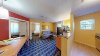 This generously sized hotel suite is designed with comfort and functionality in mind, offering a thoughtful layout across a single floor. The suite is defined by its warm, vibrant color palette — bold yellow walls in the living and sleeping areas contrast with a striking red accent wall in the dining zone, creating an energetic yet welcoming ambiance throughout.

The open living area features a plush sofa, an upholstered accent chair, and a coffee table, all set atop a richly patterned blue carpet that adds visual depth to the space. A flat-screen television is positioned for easy viewing from both the seating area and the kitchenette. Natural light streams in through multiple windows dressed with sheer curtains, providing a bright and airy atmosphere.

The fully equipped kitchen includes a full-size electric range, granite countertops, dark wood cabinetry, and ample prep space — ideal for guests on extended stays. Adjacent to the kitchen, a dedicated dining area with a wood table and seating offers a convenient space for meals.

The separate bedroom provides a private retreat, complete with a king-size bed featuring a clean white duvet, a wooden headboard, bedside tables with lamps, a work desk, and additional television. A large window with sheer curtains allows natural light to fill the room while maintaining privacy.