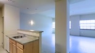 This single-floor space presents an open and functional layout that connects the kitchen and living areas seamlessly. The kitchen features a peninsula-style counter with a laminate surface and light wood cabinetry, providing practical workspace and a natural division between the cooking and living zones. A built-in sink with a standard faucet is integrated into the counter, adding everyday convenience. The flooring throughout appears to be a light-toned concrete or hard surface, lending a clean and neutral foundation to the space. Walls are finished in soft, muted tones that contribute to a calm and understated ambiance. A wall-mounted light fixture adds a warm accent to the kitchen area. Structural columns subtly define the transition between spaces without fully enclosing them, preserving the open feel. Windows positioned along the far walls allow natural light to filter in, brightening the interior and enhancing the sense of openness. The living area beyond the kitchen is generously proportioned and unfurnished, offering flexibility for a variety of layout configurations. Ceiling-mounted ventilation fixtures are also present, supporting practical air circulation throughout the space.