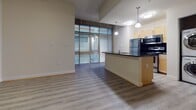 Located at 420 N 18th St in Richmond, Virginia, this single-floor unit blends modern finishes with industrial character. The open-concept living and kitchen area is anchored by durable light wood-tone flooring that flows seamlessly throughout the space. The kitchen features a dark granite countertop peninsula, warm maple cabinetry, stainless steel appliances including a refrigerator, stove, and microwave, and pendant lighting overhead. Large floor-to-ceiling windows in the living area flood the space with natural light and provide views to the exterior. A notable design feature is the exposed concrete column and raw ceiling beam in the main living area, lending an industrial loft aesthetic. Generous built-in open shelving and closet storage with wood-framed glass-paneled doors offer flexible organization options. The primary bathroom is appointed with a maple vanity, dark granite countertop, an undermount sink, a large mirror, and chrome fixtures, with tile flooring for a polished look. A second bathroom provides a tub-shower combination, chrome hardware, and clean, neutral finishes. The overall ambiance is bright, open, and contemporary, with thoughtful architectural details that give the space a distinctive character.
