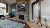 Located at 3515 S Nelson Cir in Denver, Colorado, this single-floor community space blends upscale design with a welcoming, functional layout. The grand entrance greets visitors with soaring two-story ceilings, floor-to-ceiling arched windows framed by dramatic drapery, and abundant natural light that floods the foyer. A branded entry mat and tufted navy seating create a polished first impression.

The open main living area is anchored by a striking floor-to-ceiling stacked stone fireplace topped with a solid wood mantel and a wall-mounted flat-screen television. Rich hardwood flooring transitions seamlessly into warm travertine tile throughout, adding depth and texture to the space. The lounge is furnished with a generous sectional sofa and area rug, offering a comfortable and inviting gathering environment.

An elegant arched doorway with frosted glass panels connects the main lounge to adjacent spaces, reinforcing the architectural sophistication of the interior. A dedicated wet bar area features rich knotty wood cabinetry, a stacked stone backsplash, white countertops, pendant lighting, and bar seating — ideal for entertaining. Recessed lighting and a dark-painted ceiling add a refined, intimate contrast to the bright white walls throughout. Overall, the space exudes a contemporary yet warm aesthetic, well-suited for community gatherings and social events.