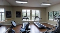 The Timber Point Fitness Center is a dedicated single-floor exercise facility designed to support an active lifestyle. The space features warm hardwood flooring throughout, which adds a clean and polished aesthetic to the room. Overhead fluorescent lighting ensures the area is well-lit, complemented by natural light streaming through a large multi-pane window that offers views of the surrounding greenery.

The fitness center is equipped with a selection of cardio machines, including multiple treadmills positioned near the window and additional equipment such as a recumbent bike and step machine. The layout is thoughtfully arranged to provide ample space between machines, allowing for comfortable and safe use. Neutral-toned walls create a calm, focused environment conducive to exercise.

A posted Fitness Center Policy sign is prominently displayed, keeping members informed of facility rules and guidelines. The room's design prioritizes functionality while maintaining a tidy, welcoming atmosphere. Clean lines, practical fixtures, and a straightforward layout make this fitness center a convenient and well-maintained amenity for its users.