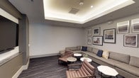 This theater space is thoughtfully designed to blend comfortable viewing with sophisticated lounge-style aesthetics. The room centers around a large flat-screen television mounted within a recessed niche, accompanied by a sleek soundbar and a stone-finish ledge beneath, creating a polished focal point for entertainment.

The seating arrangement is both generous and well-considered, featuring a substantial gray sectional sofa accented with navy, white, and yellow throw pillows, complemented by a set of rich brown leather lounge chairs with wooden frames. Hexagonal and round white marble-top side tables are positioned throughout for added functionality and visual interest.

A standout architectural feature is the tray ceiling with integrated ambient cove lighting, which casts a warm, even glow across the room, enhancing its inviting atmosphere. The dark, patterned carpet with subtle metallic veining adds depth and texture underfoot.

One wall serves as a curated gallery, displaying an eclectic collection of framed artwork in varying sizes, including nature-inspired prints of horses, deer, feathers, and landscapes, adding a refined, artistic character to the space. A sliding glass door connects the theater to an adjacent, brighter common area, offering seamless flow between spaces.