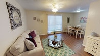 This single-floor space centers around a well-appointed living area that blends comfort with style. Dark hardwood flooring runs throughout, providing a rich, grounding contrast to the light-toned walls. A textured ceiling adds subtle character overhead, while a ceiling fan with an integrated light fixture ensures year-round comfort and practical illumination.

Natural light filters through a large window fitted with white horizontal blinds, brightening the room and creating an open, airy feel. The living area is thoughtfully arranged, anchored by a tufted sofa and an accent chair that invite relaxation. A round wooden coffee table sits atop a plush white area rug, adding warmth and a natural element to the space.

A bold, large-scale abstract painting in yellows, blues, and blacks serves as a striking focal point on one wall, adding artistic character. Lush potted plants positioned throughout the room introduce a refreshing natural element. A tripod floor lamp provides additional ambient lighting, rounding out a space that balances everyday livability with thoughtful design details.