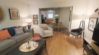 This single-floor residence offers a thoughtfully designed open-concept layout that seamlessly connects the living, dining, and kitchen areas into one cohesive and inviting space. Warm hardwood flooring flows throughout, tying each area together and lending a sense of continuity and warmth.

The living area is comfortably appointed with a plush sofa, a cushioned accent chair, and a dark coffee table accessorized with a decorative tray — creating a relaxed yet polished setting for everyday living and entertaining. Framed artwork adorns the walls, adding character and a personal touch.

The dining area is situated just beyond, accommodating a round wood table with seating for multiple guests. A striking floral arrangement serves as a natural centerpiece, adding a fresh, elegant accent to the space.

Floor-to-ceiling drapery along one wall softens the room and provides privacy while contributing to the overall refined aesthetic. Track lighting overhead ensures the space is well-lit throughout.

The kitchen, visible through an open passthrough, features warm wood cabinetry, modern appliances including a full-size refrigerator, and ample counter space — ideal for everyday meal preparation. A window above the sink allows natural light to brighten the kitchen area. Overall, the space offers a functional, comfortable, and stylishly curated living environment.