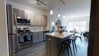 This single-floor residence showcases a thoughtfully designed open-concept layout that seamlessly connects the kitchen, dining, and living areas into one cohesive, airy space. The kitchen is outfitted with stainless steel appliances, including a gas range and built-in microwave, complemented by warm-toned flat-front cabinetry that offers ample storage. A sleek countertop runs the length of the kitchen, providing generous workspace, while a central island with a light quartz surface and dark wood bar stools serves as a natural gathering point. Elegant pendant lights hang above the island, adding a refined touch to the space. Track lighting along the ceiling ensures the area is well-lit throughout. The open dining area flows naturally from the kitchen, bathed in natural light streaming through large windows that brighten the entire living space. Wide-plank hardwood-style flooring in a cool grey tone extends throughout, tying the rooms together with a clean, modern aesthetic. The overall ambiance is contemporary and inviting, with a neutral color palette and high-quality finishes that create a sophisticated yet functional living environment.