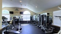 This fitness center offers a thoughtfully designed workout space beneath dramatic vaulted ceilings that lend an open, airy feel to the room. A prominent arched window serves as a focal point, allowing natural light to flood the interior and creating a bright, energizing atmosphere. Recessed lighting and a ceiling fan provide additional comfort during workouts.

The space is outfitted with a comprehensive selection of exercise equipment, including a multi-station cable machine, weight benches, free weights, dumbbells, and various strength training apparatus arranged to maximize usable floor space. Rubber flooring throughout the workout area provides both safety and durability underfoot.

Wall-mounted mirrors along one side of the room visually expand the space while allowing users to monitor their form during exercise. A wall-mounted television adds an entertainment element for those looking to stay engaged during longer sessions. A branded logo on the wall indicates this is an amenity space associated with a residential community. The overall design balances functionality with comfort, making it a practical and accessible facility for a variety of fitness routines.