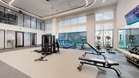 Located at 1133 Harding Place in Charlotte's vibrant 28204 corridor, this contemporary fitness center is thoughtfully designed to support a full spectrum of workout routines. The main exercise floor boasts soaring double-height ceilings and expansive floor-to-ceiling windows that flood the space with natural light and offer views of the surrounding streetscape. Curved LED lighting traces the ceiling, lending the area a sleek, modern ambiance. The flooring transitions between light-toned performance surfaces and darker carpet zones, delineating different workout areas with purpose and style. A comprehensive selection of Precor cardio machines—including treadmills, stationary bikes, and rowing equipment—lines the windowed perimeter, while a multi-station cable machine and adjustable benches anchor the central strength-training area. A dedicated functional training zone, finished with bold green accent walls and full-length mirrors, is equipped with TRX suspension systems, battle ropes, medicine balls, and turf training lanes. A separate studio room features herringbone hardwood flooring, exposed wooden ceiling beams, warm pendant lighting, full mirrored walls, and space for yoga, stretching, or group classes. The entrance area showcases geometric wall art, open wood shelving, and glass-panel doors, creating a welcoming and well-organized arrival experience.