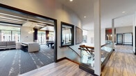 Located at 2007 Knoll Top Ln in Franklin, TN, this contemporary amenity facility delivers an impressive array of thoughtfully designed spaces. The grand multi-level interior is anchored by a soaring atrium lobby with floor-to-ceiling windows, a striking chandelier, and an elegant curved staircase with glass balustrades and a warm wood handrail. Wide-plank wood-look flooring and curated artwork flow throughout the corridors, creating a polished, gallery-like atmosphere. The game room is a standout feature, equipped with a billiards table, shuffleboard, a large-format wall-mounted TV, and a decorative guitar art installation—nodding to the region's musical heritage. An adjacent lounge area offers built-in upholstered bench seating, geometric side tables, and patterned carpeting beneath a dramatic dark coffered ceiling with ample natural light from surrounding windows. The fitness center is well-appointed with strength training equipment, rowing machines, punching bags, and floor-to-ceiling mirrors, all set against a bold geometric carpet. A dedicated yoga and stretching studio with bamboo flooring and mirrored walls completes the wellness offerings. Black-framed glass partition doors and high ceilings throughout contribute to a modern, open, and sophisticated environment.