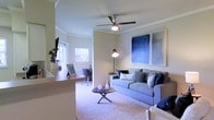 This single-floor residence offers a thoughtfully designed open-concept layout that maximizes both functionality and comfort. The living area serves as the central hub of the space, anchored by plush wall-to-wall carpeting and a generously sized sofa dressed in cool blue and grey tones, complemented by coordinating accent pillows. A round wooden coffee table adds warmth and serves as a natural focal point.

Crown molding runs along the perimeter of the ceiling, lending a polished architectural detail to the room. A ceiling fan with an integrated light fixture provides both ventilation and ambient illumination, while a freestanding arc floor lamp offers additional task lighting. Large framed blue abstract artwork adorns the wall, reinforcing the cohesive cool-toned color palette throughout.

The space transitions seamlessly into a dedicated workspace area, visible in the background with a desk and chair, as well as a kitchen counter or bar overhang that defines the boundary between living and kitchen zones. Natural light filters in through a window with white blinds, brightening the overall atmosphere. A wall-mounted television and indoor greenery complete the inviting, well-balanced aesthetic of this versatile living space.