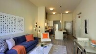 This stylish single-floor space showcases a thoughtfully designed open-concept layout where the living area and kitchen flow naturally into one another. The living room is anchored by a bold navy blue sofa accented with vibrant orange throw pillows, complemented by a sleek white accent chair, a geometric coffee table, and a patterned area rug. A striking multi-arm sculptural floor lamp adds an artistic focal point to the space. The soaring ceiling height creates an airy, expansive atmosphere that enhances the sense of openness throughout.

The kitchen is a standout feature, boasting dark cabinetry contrasted against light countertops, a stainless steel refrigerator, a tile backsplash, and a peninsula with bar seating — ideal for casual dining or entertaining. Pendant lighting above the peninsula adds warm, focused illumination.

Durable wood-look flooring runs throughout, tying all areas together cohesively. Flush-mount ceiling lights provide consistent, even lighting across the space. The overall aesthetic is clean and contemporary, with a neutral warm color palette punctuated by carefully chosen accent colors. The combination of functional design and modern finishes makes this a well-appointed and inviting space.
