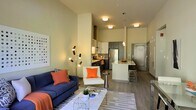 This stylish single-floor space showcases a thoughtfully designed open-concept layout where the living area and kitchen flow naturally into one another. The living room is anchored by a bold navy blue sofa accented with vibrant orange throw pillows, complemented by a sleek white accent chair, a geometric coffee table, and a patterned area rug. A striking multi-arm sculptural floor lamp adds an artistic focal point to the space. The soaring ceiling height creates an airy, expansive atmosphere that enhances the sense of openness throughout.

The kitchen is a standout feature, boasting dark cabinetry contrasted against light countertops, a stainless steel refrigerator, a tile backsplash, and a peninsula with bar seating — ideal for casual dining or entertaining. Pendant lighting above the peninsula adds warm, focused illumination.

Durable wood-look flooring runs throughout, tying all areas together cohesively. Flush-mount ceiling lights provide consistent, even lighting across the space. The overall aesthetic is clean and contemporary, with a neutral warm color palette punctuated by carefully chosen accent colors. The combination of functional design and modern finishes makes this a well-appointed and inviting space.
